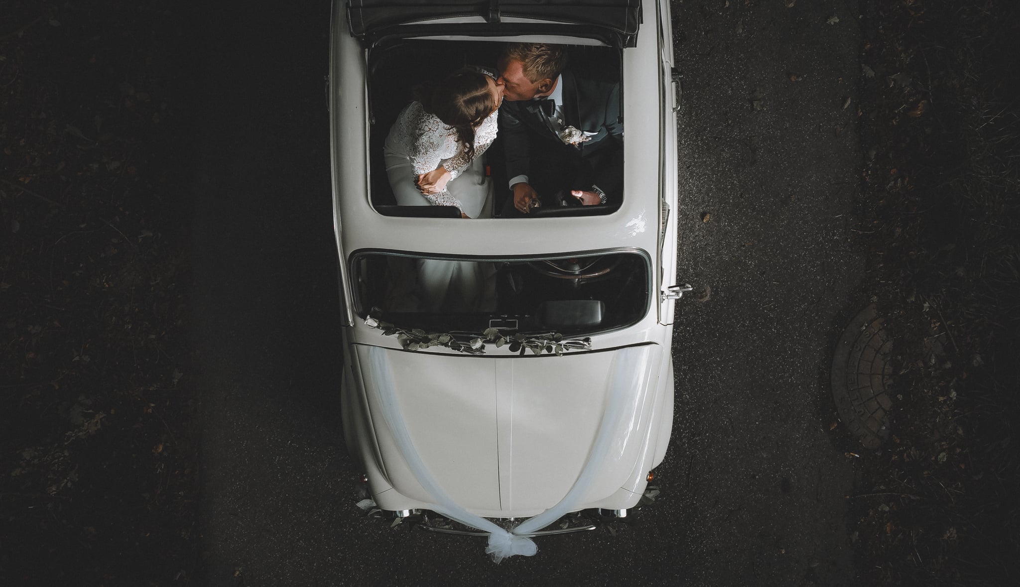 Dronebillede taget af et smukt brudepar, der sidder i en hvid Fiat 500 med soltag. Den er pyntet med tyl og blomster. Soltaget er åbnet og med dronen har jeg fanget dem, hvor de sidder og kysser hinanden set oppefra.